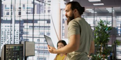 Cheerful admin holding laptop chatting with colleagues while working with machine learning tech. Man transforming data into predictive models using notebook and talking with coworkers, camera A