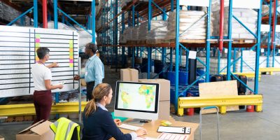 High angle view of mature female manager working on computer while coworkers discussing over whiteboard in warehouse. This is a freight transportation and distribution warehouse. Industrial and industrial workers concept