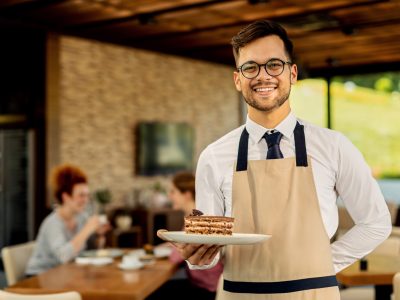We serve the best cakes! Happy waiter with slice of cake standing in a cafe and looking at camera. There are people in the background.