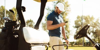 Young man using mobile phone while standing at a golf cart on a field